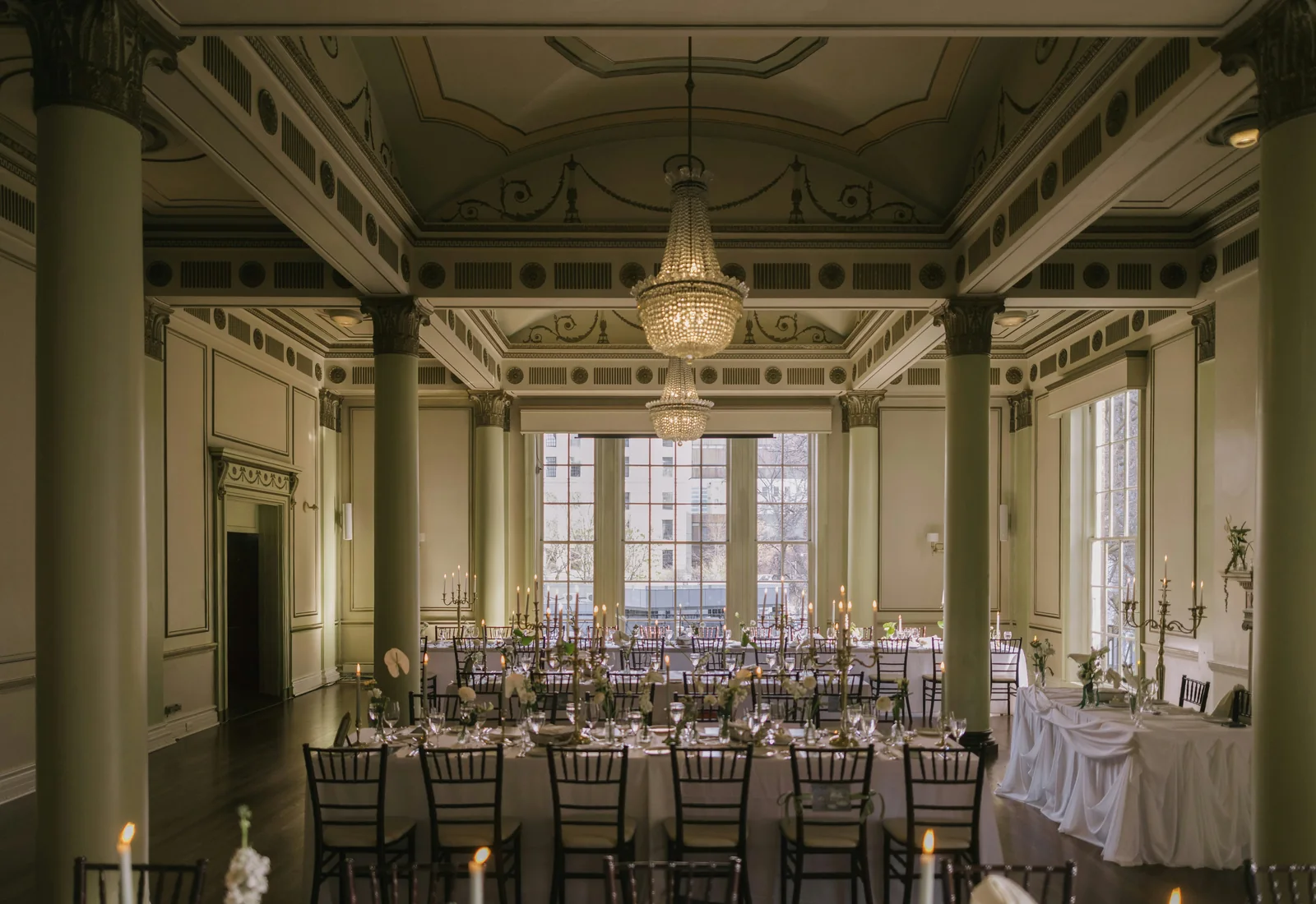 An ornate columned banquet hall set with long dining tables, candelabras and a crystal chandelier, representative of a period Viennese dinner room